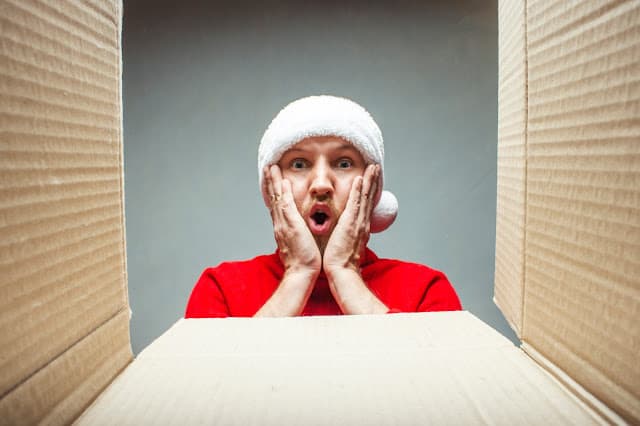 Man with a surprised face in a Santa Claus hat and a red sweater looks into a large, empty cardboard box.
