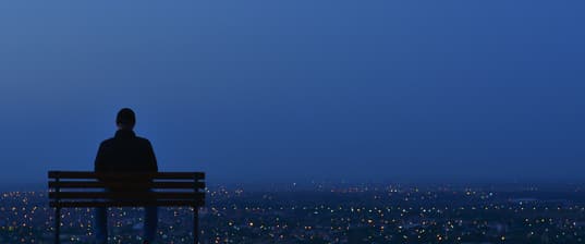 Silhouette of a person sitting on a park bench looking out over city lights.